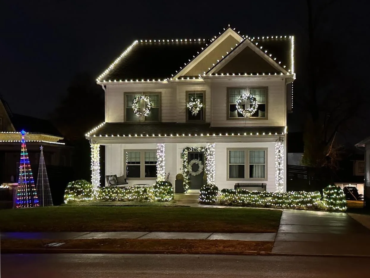 Two-story home with white Christmas lights along rooflines, porch columns and shrubs