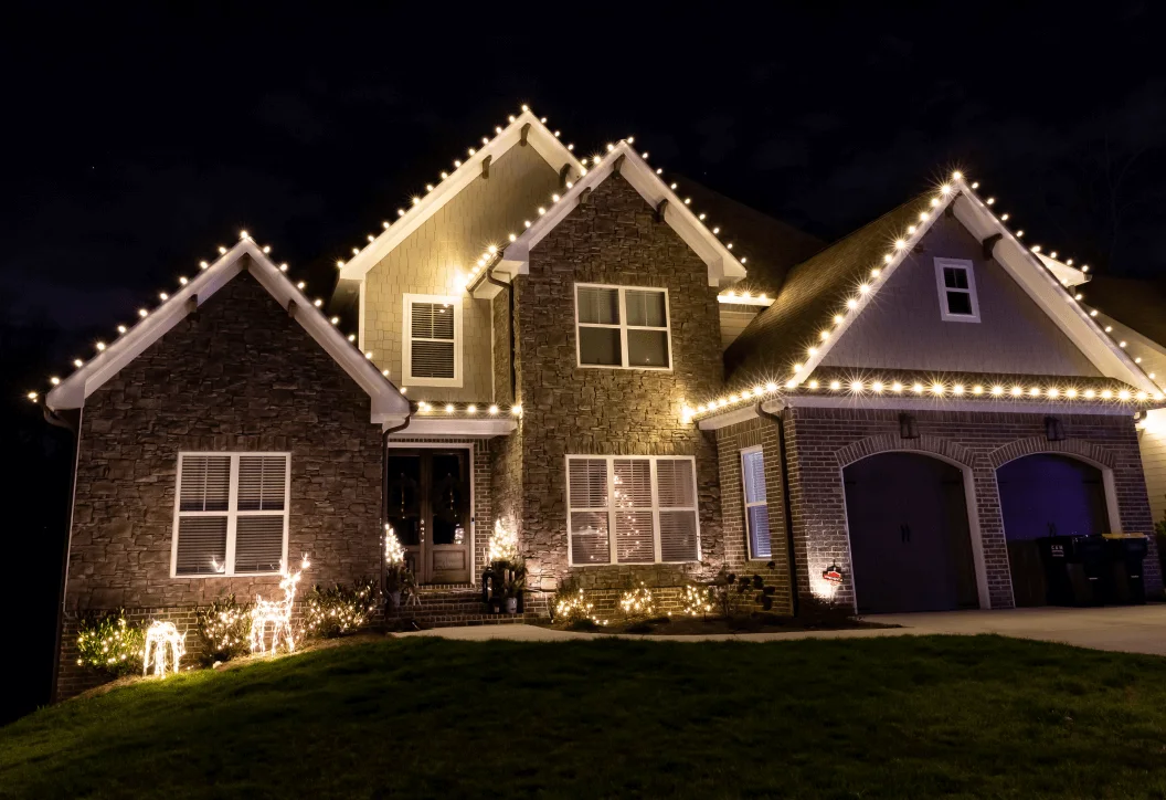 Stone-front home with warm white Christmas lights outlining the gables and illuminated lawn decorations
